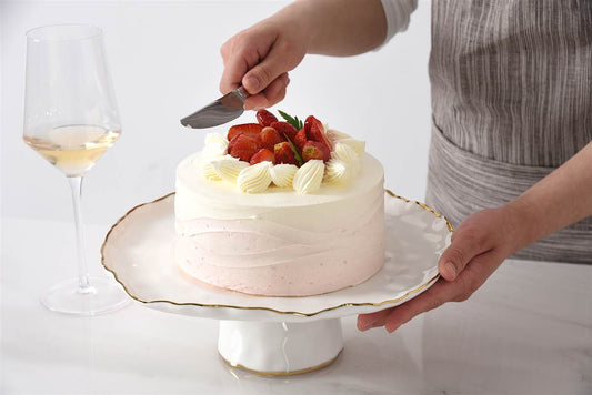 Person cutting a cake with strawberries on a white plate, with a glass of wine in the background.