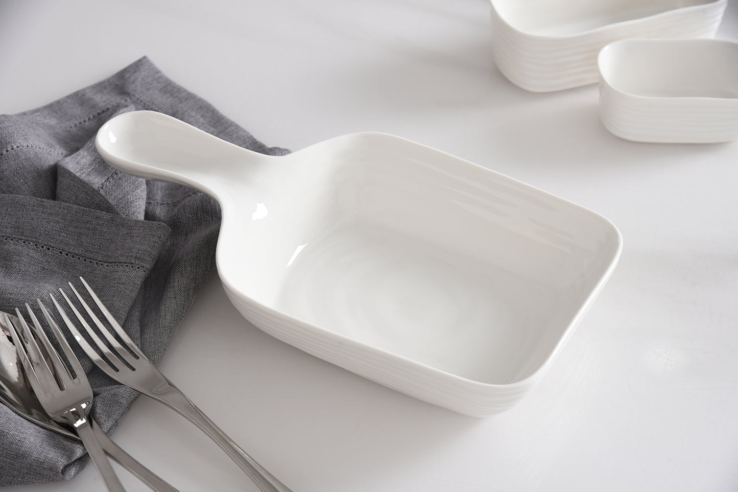 White ceramic serving spoon and dish on a white surface with gray napkins and silver forks.