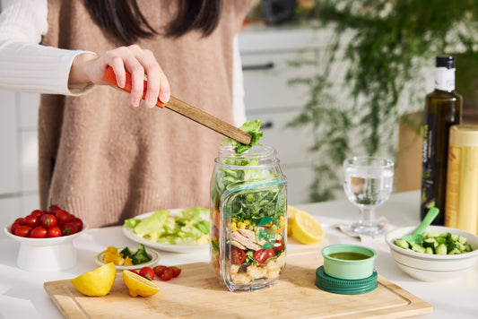 Person preparing a salad in a jar with ingredients on a kitchen counter.