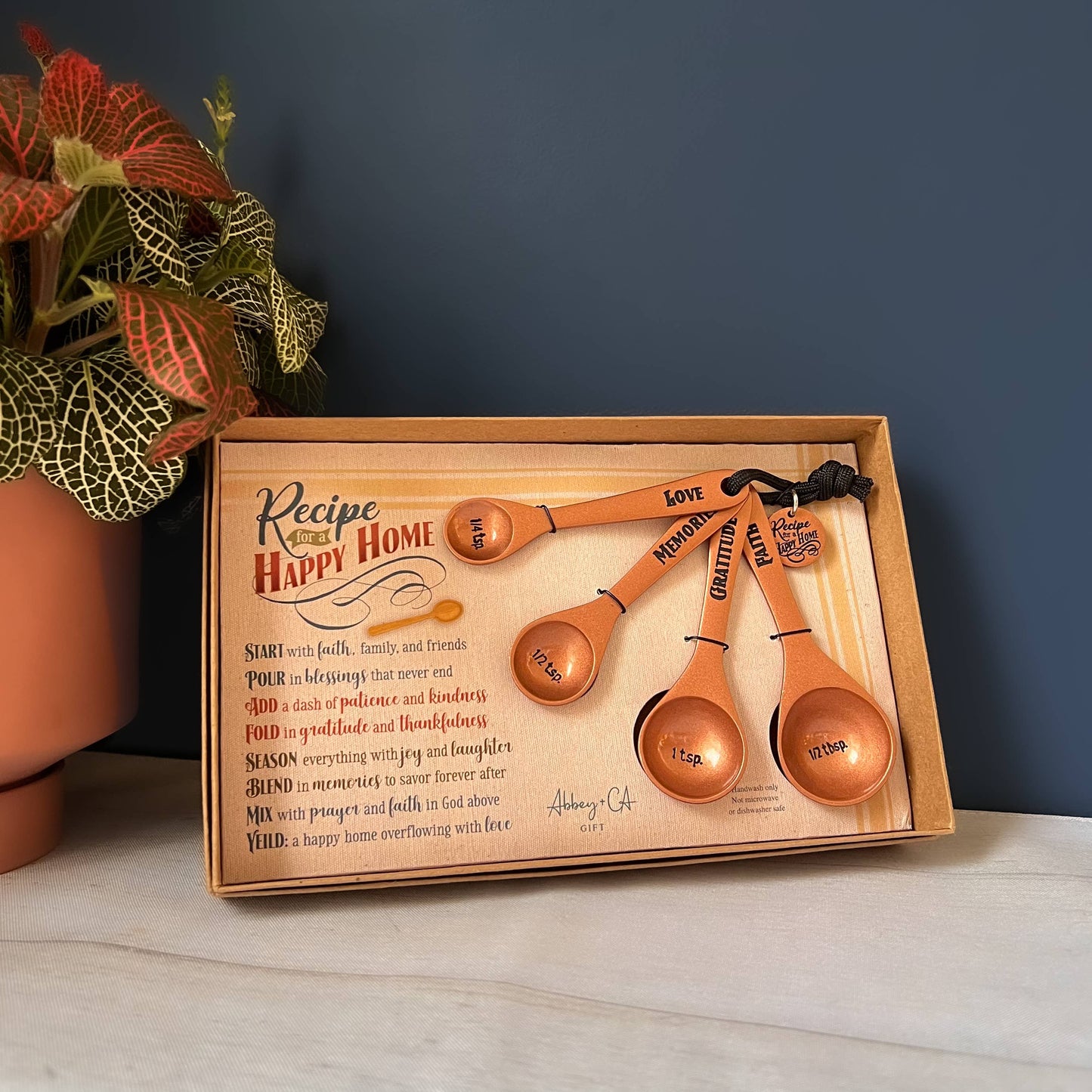 Set of copper measuring spoons in a wooden box with 'Recipe for a Happy Home' text, on a neutral background.