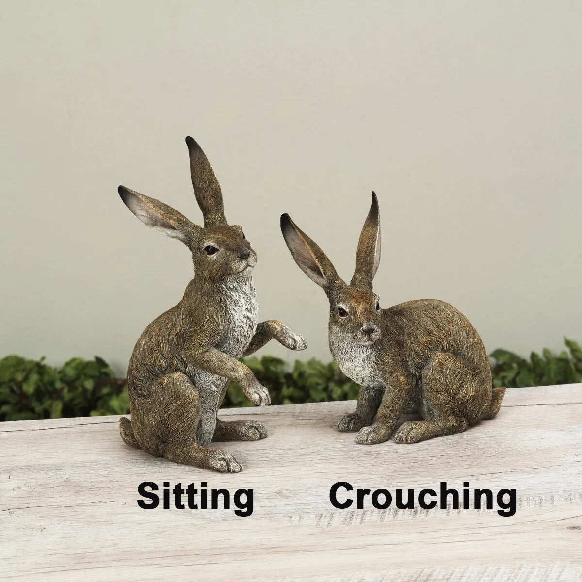 Two rabbit statues, one sitting and one crouching, on a wooden surface with a neutral background.