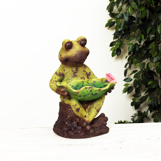 Frog statue holding a pink flower with green leaves in the background