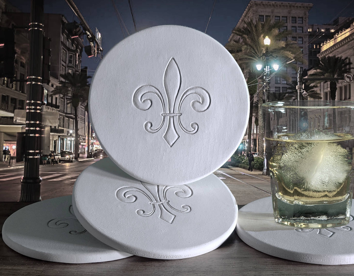 White coasters with fleur-de-lis design on a table with a glass of water in an urban night setting.