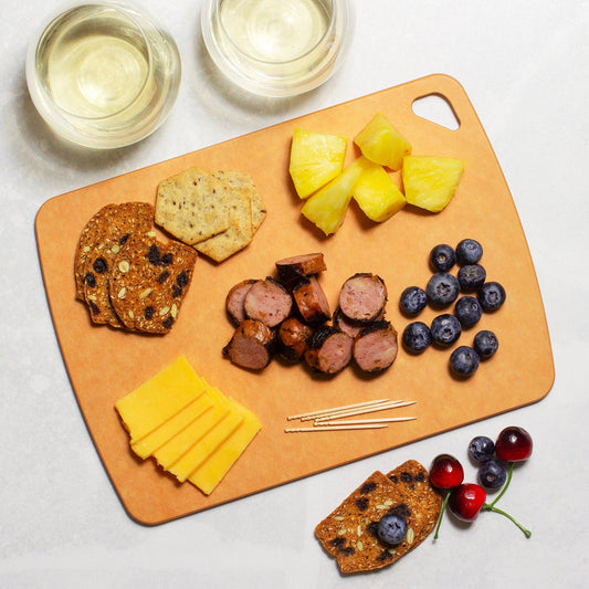 Wooden cutting board with assorted foods including sausages, cheese, crackers, and fruit on a light background.