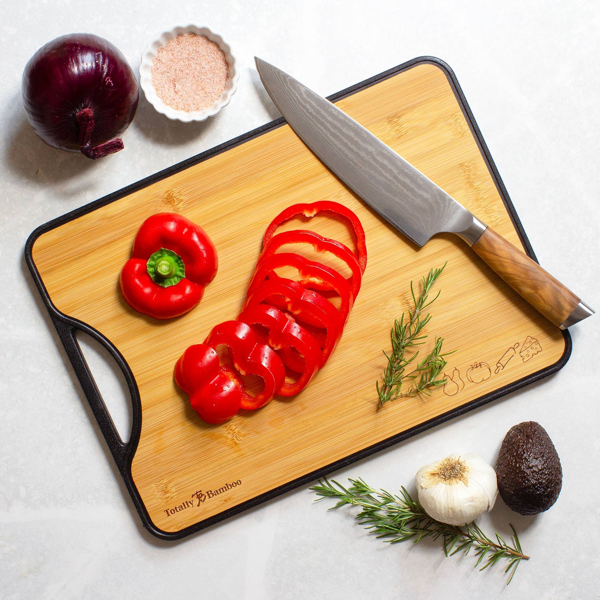 Wooden cutting board with sliced red pepper, knife, and various vegetables on a white surface.