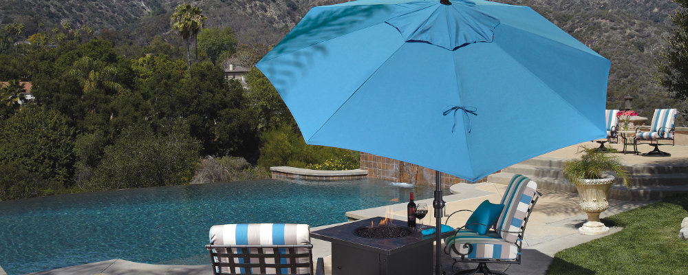 Blue patio umbrella over a fire pit with a pool and garden in the background