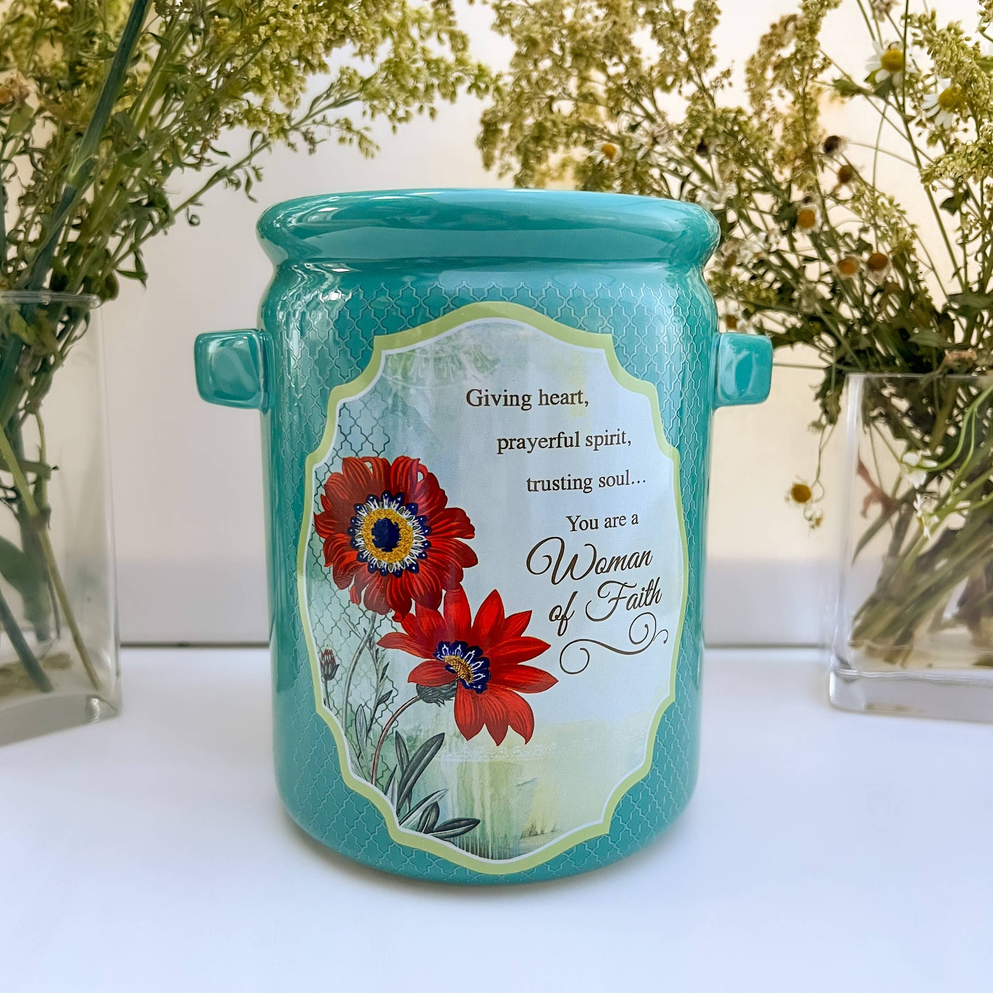 Turquoise jar with floral design and text on a white surface with plants in the background