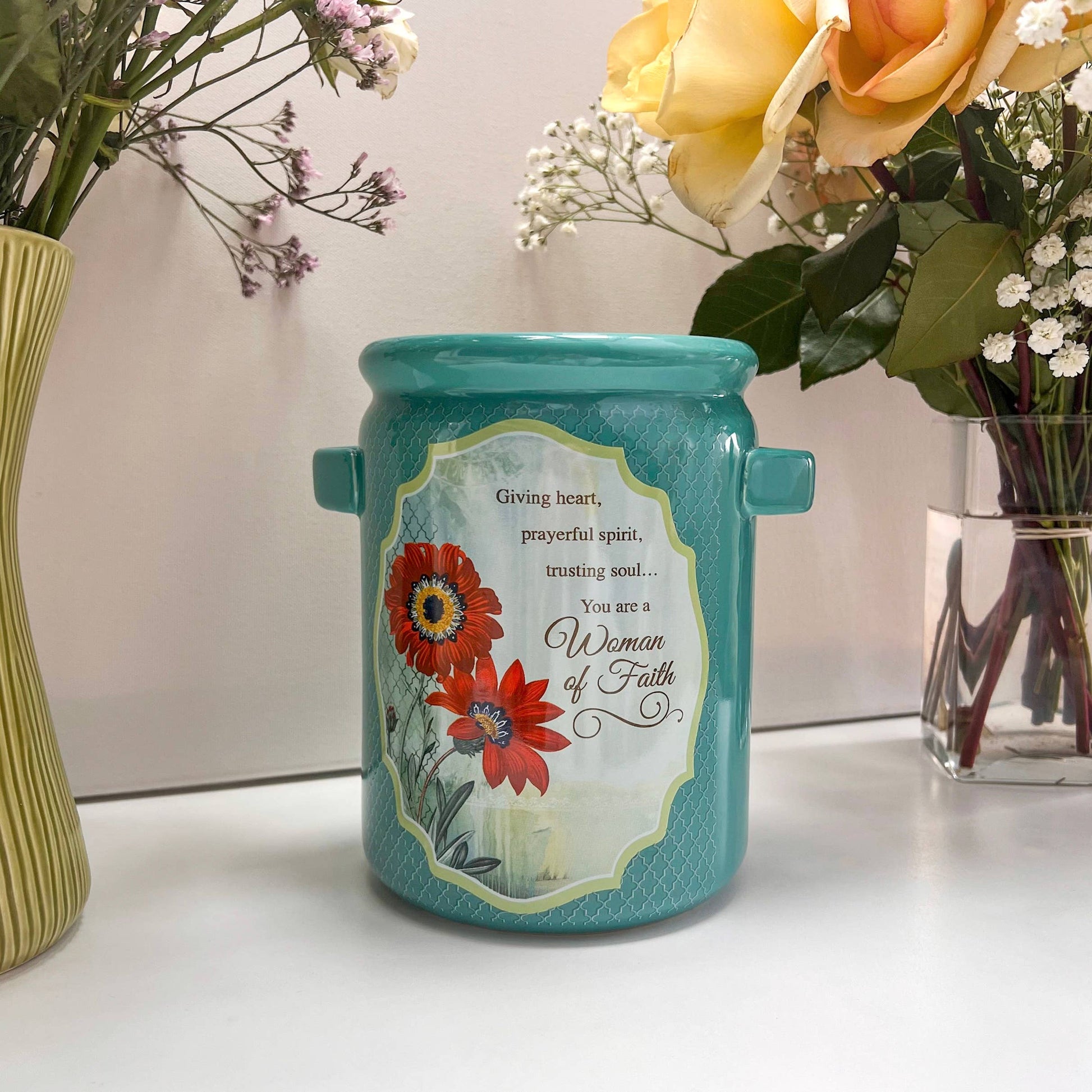 Turquoise ceramic jar with floral design and text, placed on a surface with flowers in the background.