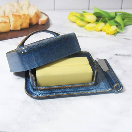 A blue ceramic butter dish with a handle, shown open with a stick of butter inside and a knife resting on the side, set on a white marble surface with bread and yellow tulips in the background.