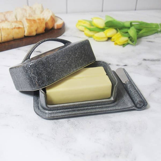 A dark gray speckled ceramic butter dish with a handle and matching knife, shown with a stick of butter inside. The scene is set on a white marble countertop with a blurred background showing sliced bread on a wooden board and yellow tulips.