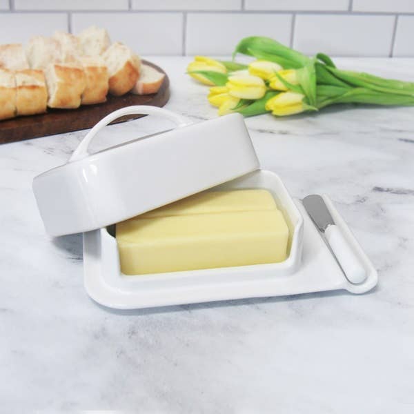 A white ceramic butter dish with a matching lid, shown with a stick of butter inside and a small white-handled butter knife on a marble countertop. Yellow tulips and sliced bread are blurred in the background.