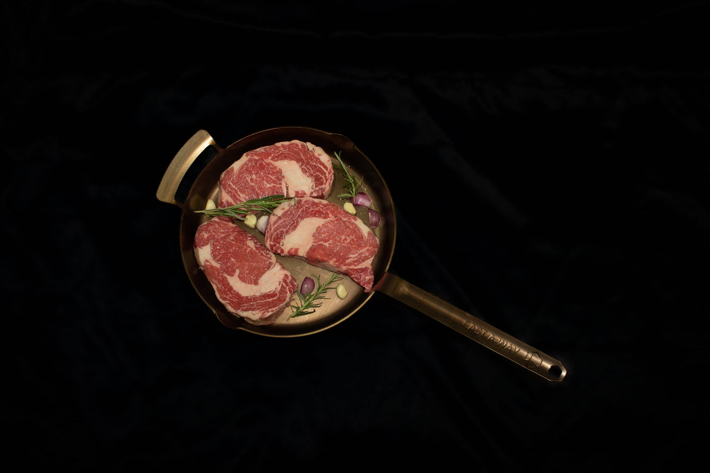 A top-down view of a small frying pan with a long handle, containing two raw marbled beef steaks topped with herbs, set against a dark black background.