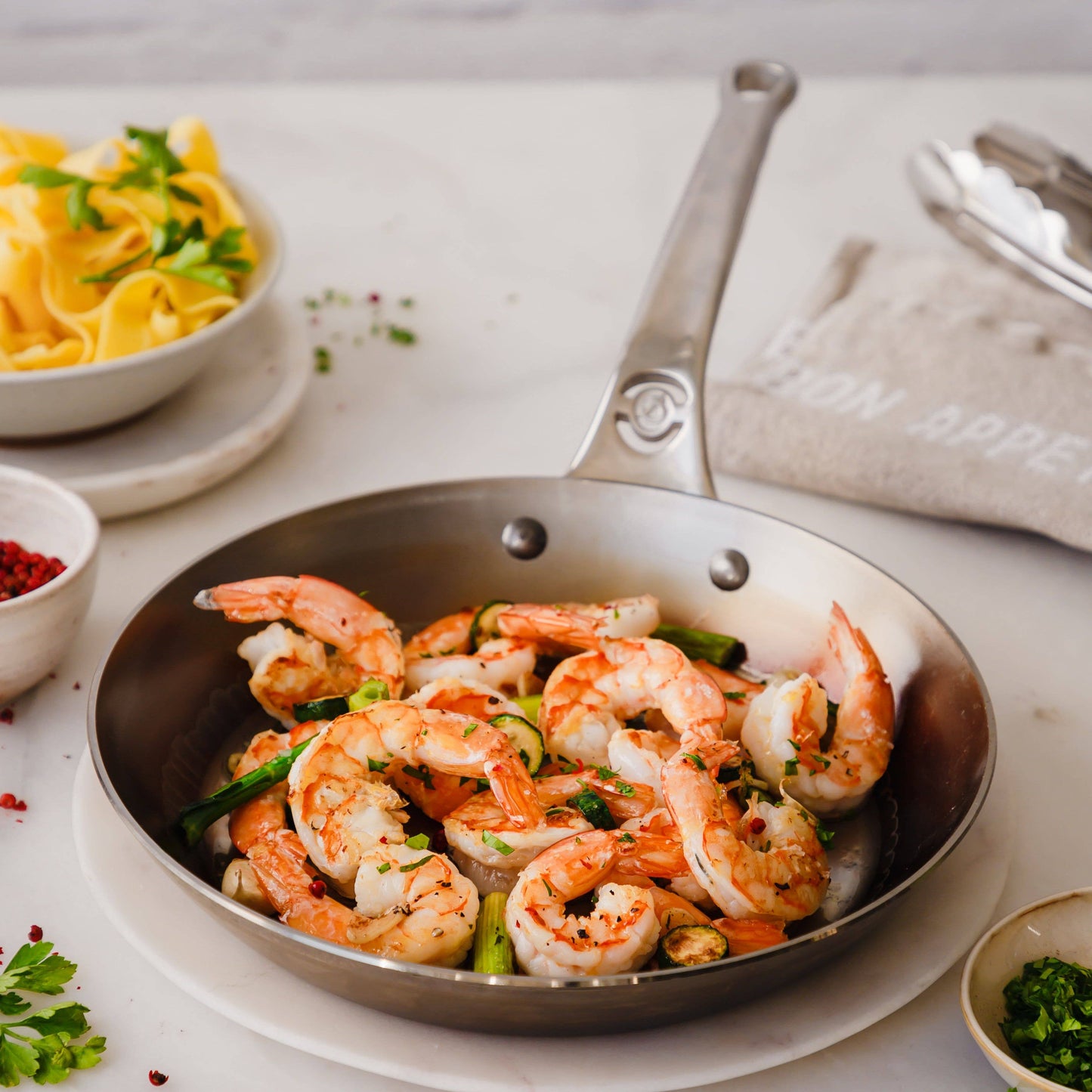 Frying pan with shrimp and vegetables on a table with pasta and herbs.