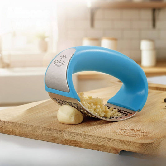 Blue kitchen garlic tool on a wooden cutting board with garlic being crushed.