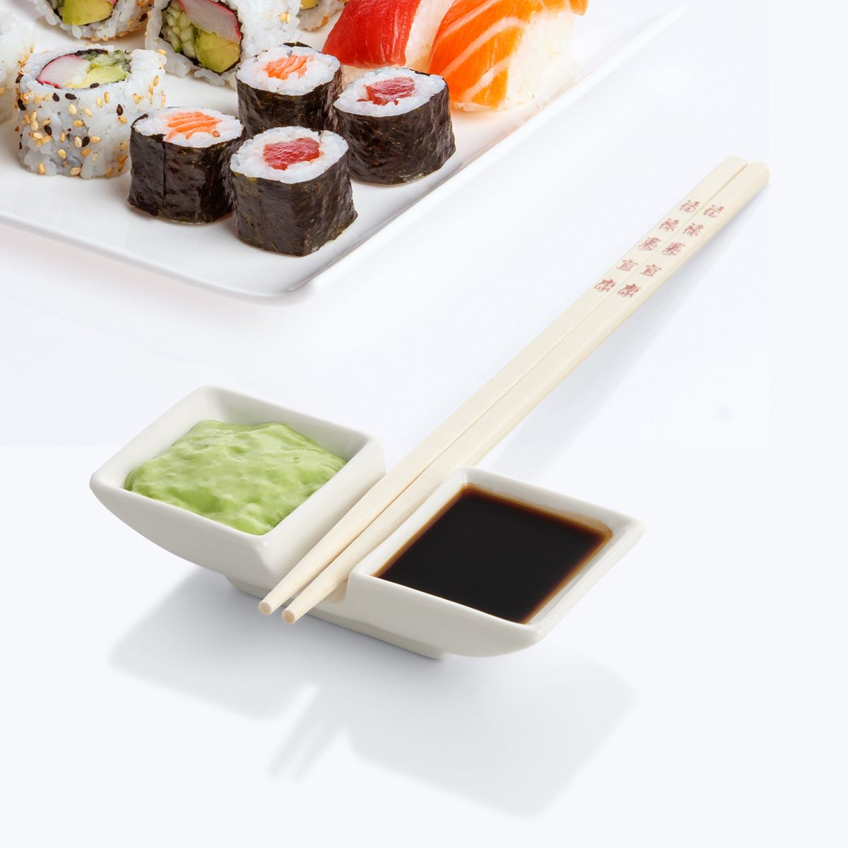 Sushi set with sushi rolls, wasabi, soy sauce, and chopsticks resting on a ceramic dish on a white surface.