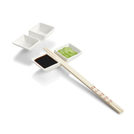 Set of chopsticks resting on a ceramic dish with soy sauce and wasabi on a white background