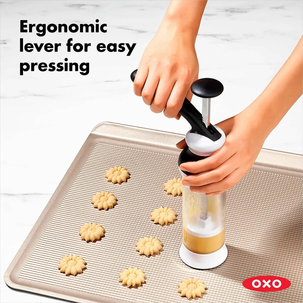OXO cookie press being used to make cookies on a baking sheet with text 'Ergonomic lever for easy pressing'.