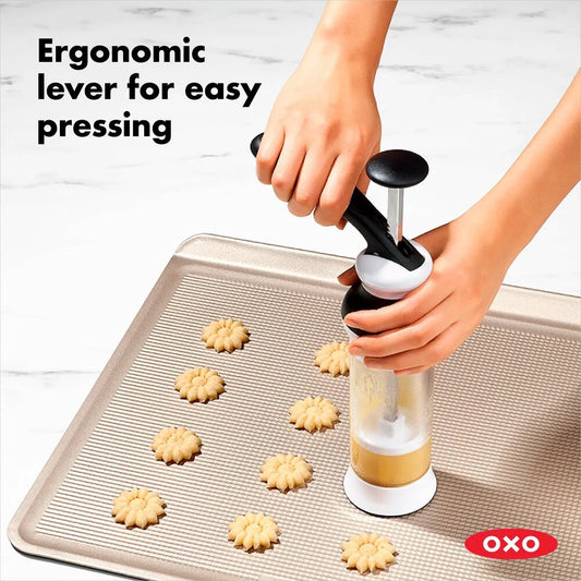 OXO cookie press being used to make cookies on a baking sheet with text 'Ergonomic lever for easy pressing'.