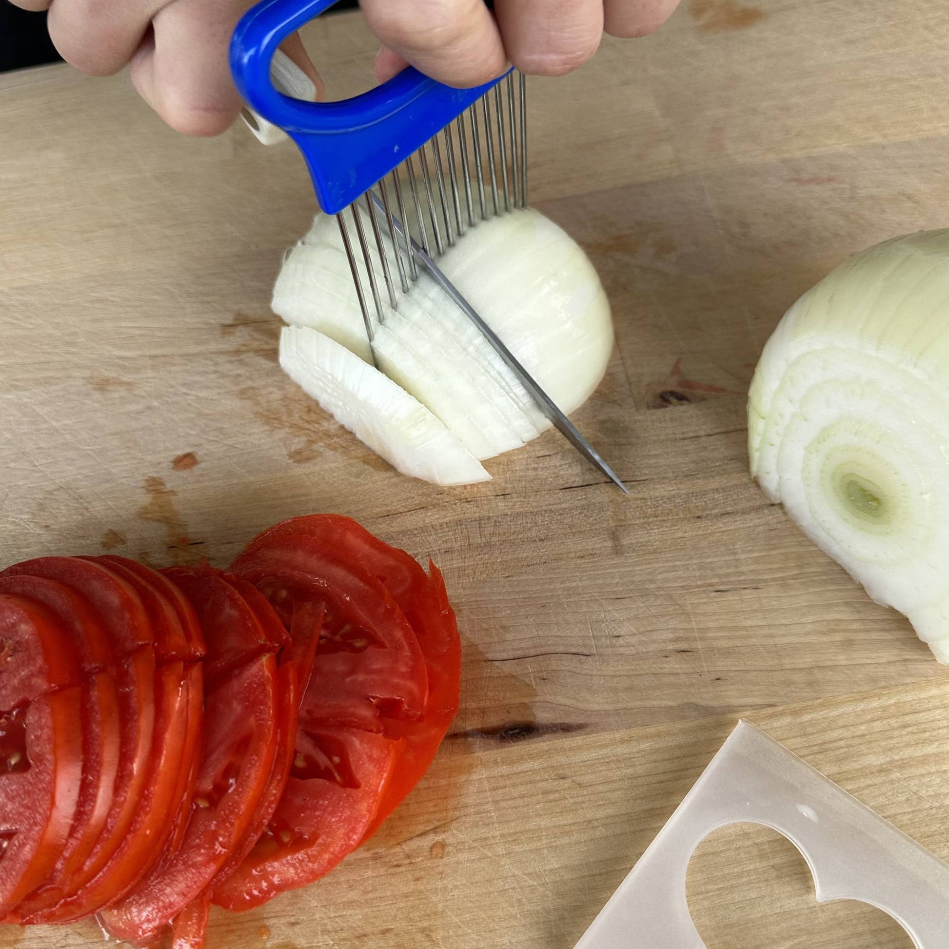Onion being sliced with a blue hand-held slicer on a wooden cutting board with sliced tomatoes.