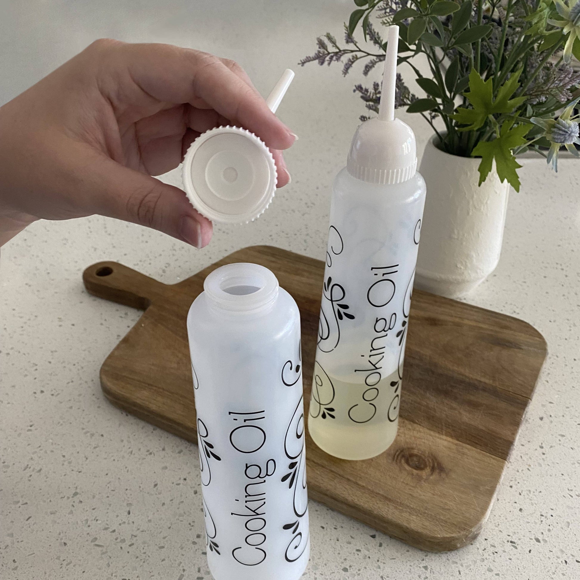 Two bottles labeled 'Cooking Oil' on a wooden cutting board with a hand holding a white cap.