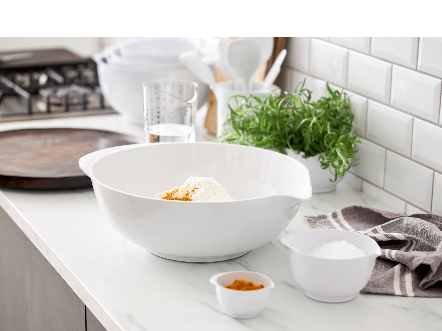 A large white Rosti mixing bowl with side handles sits on a white kitchen countertop, containing flour and a small amount of orange spice. Smaller white bowls, a measuring cup, a green herb plant, and a white subway-tile backsplash are visible in the background.
