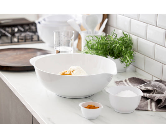 A large white Rosti mixing bowl with side handles sits on a white kitchen countertop, containing flour and a small amount of orange spice. Smaller white bowls, a measuring cup, a green herb plant, and a white subway-tile backsplash are visible in the background.