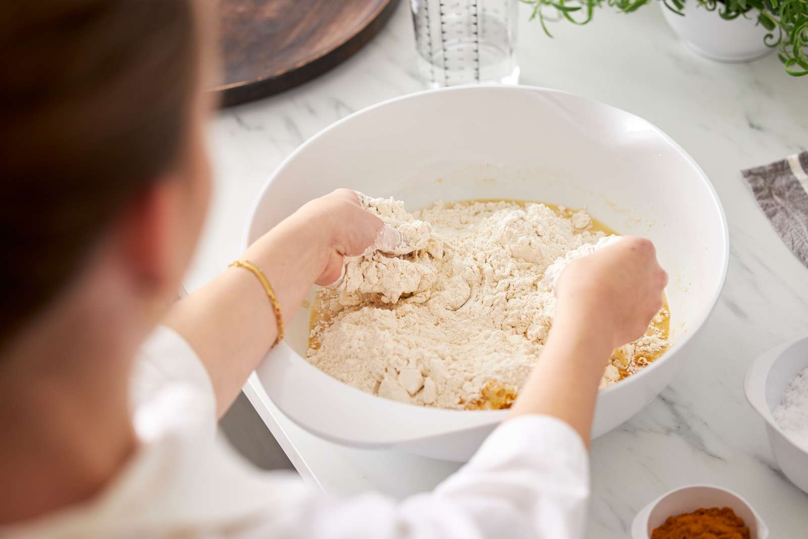 A person mixing dry ingredients, likely flour, into a wet batter in a large white bowl on a white marble countertop.