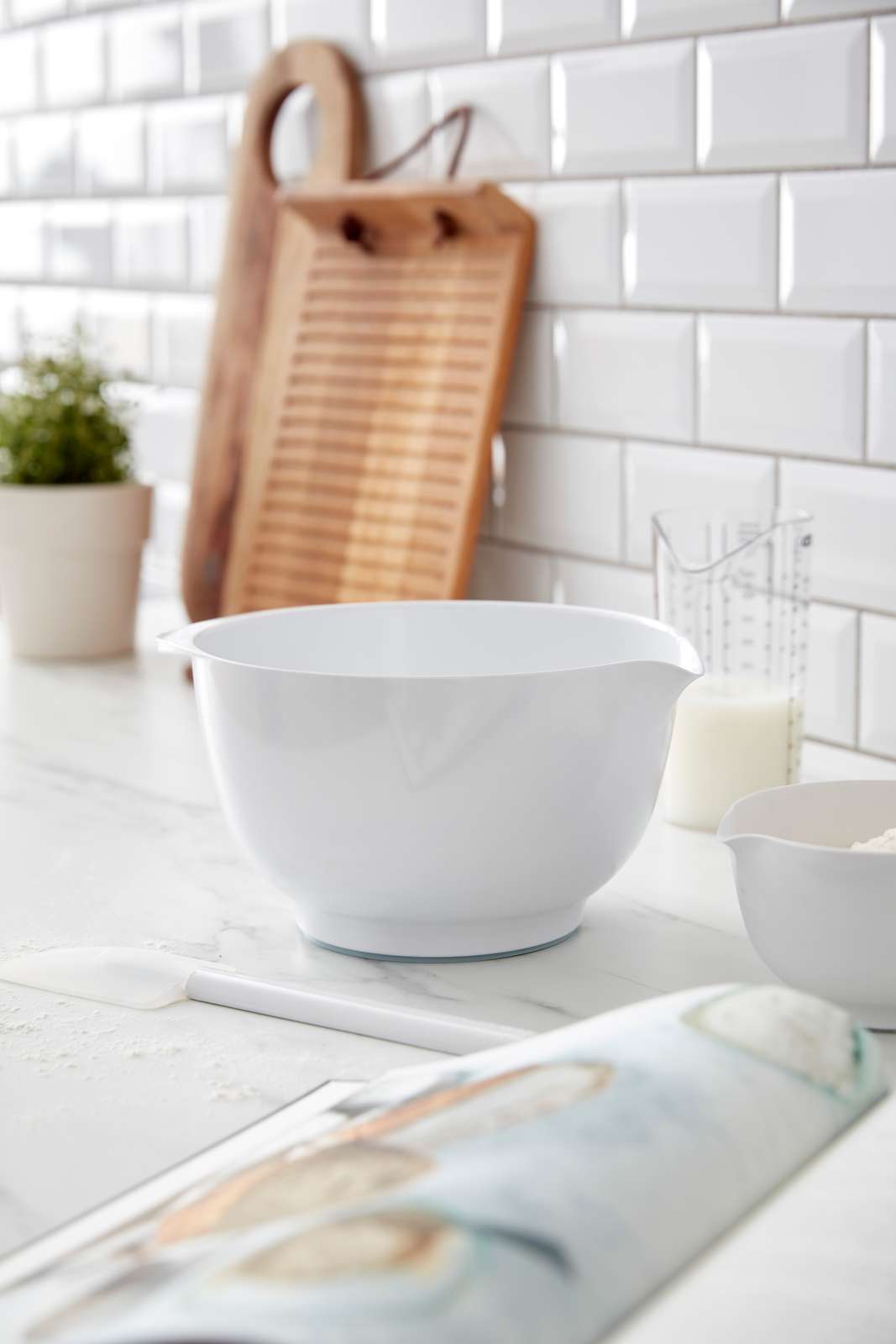 A white Rosti Margrethe mixing bowl with a pouring spout sits on a light marble countertop in a bright kitchen setting with white subway tiles, a wooden cutting board, and baking ingredients.