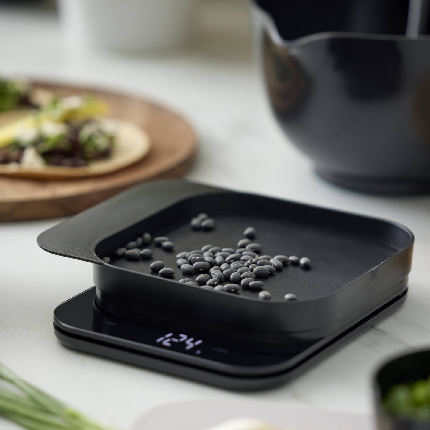 A black digital kitchen scale with a square weighing tray, shown in a kitchen setting with beans on the tray and blurred food items in the background.