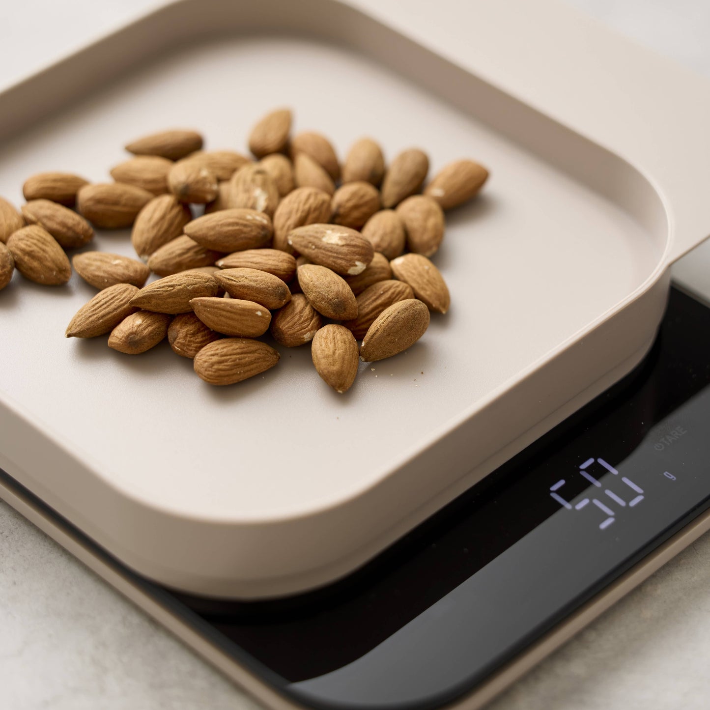 A pile of whole almonds sits on a light beige digital kitchen scale with a black display, photographed in a clean close-up kitchen setting.