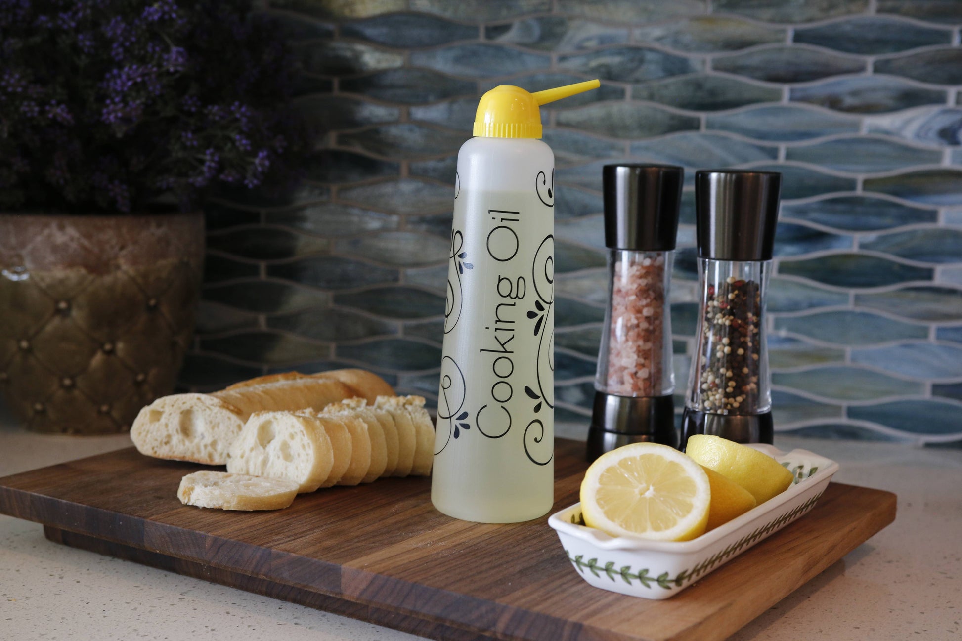 yellow Butterie oil dispenser on a cutting board with bread and lemons, against a tiled wall background.